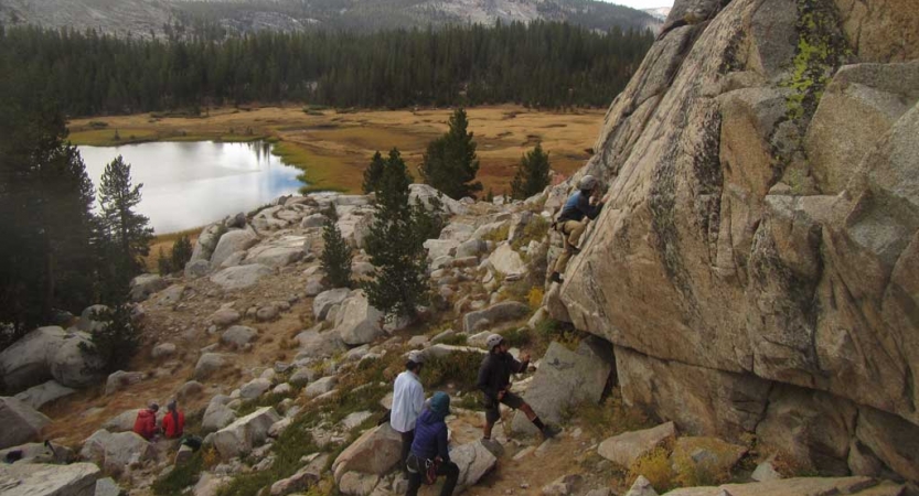 A group of people rock climb on a steep rock wall in the foreground, with an alpine meadow, lake and evergreen trees appear in the background. 